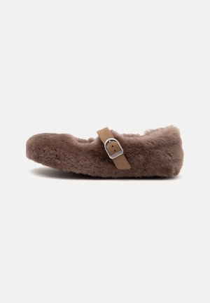 Brun, blød flad sko med en lysebrun spænde-rem over toppen, vist fra siden på en ensartet lys baggrund.