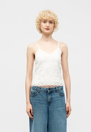 Νεαρός με κοντά, σγουρά ξανθά μαλλιά, φοράει άσπλο δαντελένιο καμιζολάκι και μπλε τζιν ψηλόμεσο, στέκεται μπροστά σε ένα μονόχρωμο φόντο.