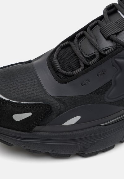 Chaussure de sport noire avec une tige multi-texturée, comprenant des composants en maille et en caoutchouc, renfort à l'avant du pied, et accents réfléchissants. Semelle épaise à crampons.