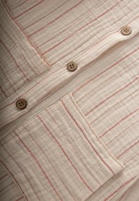 Tejido ligero de color crema con rayas rojas. Presenta botones de madera y un bolsillo en el pecho, mostrando un diseño texturizado y arrugado.