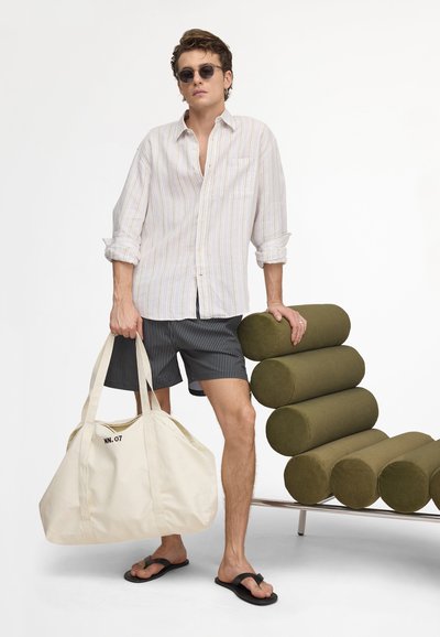 Homme en chemise rayée et short, portant des lunettes de soleil et des tongs, tenant un sac polochon blanc, appuyé sur une chaise cylindrique coussin olive.