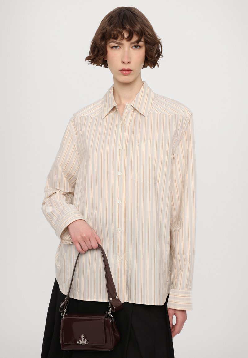 Camisa de botones beige y crema con ligeras rayas y un bolsillo en el pecho. Modelo sosteniendo un pequeño bolso oscuro rojo brillante con un cierre.