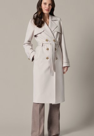 Kobieta nosząca beżowy dwurzędowy płaszcz trencz z paskiem na taupe szerokich spodniach, stojąca na tle jednolitego, neutralnego tła.