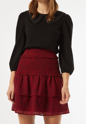 Kvinde iført en sort bluse med ballonærmer og en flæsekantet krave, kombineret med et rødt lagdelt skørt med smock-facon i taljen.