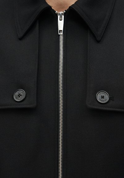 Κοντινή λήψη μαύρου μπουφάν με ασημένιο φερμουάρ, το οποίο είναι τοποθετημένο κεντρικά ανάμεσα σε δύο μαύρα κουμπιά σε πατιλέτες κοντά στον γιακά.