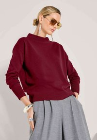 Vrouw in een bordeauxrode trui, grijze plooienbroek, grote oorbellen, een chunky armband, een oversized ring en gele zonnebril.