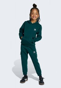 Sötétzöld kapucnis pulóver kenguru zsebbel és fehér logóval, hozzá illő terep nadrággal párosítva. A modell fekete, fehér részletekkel díszített tornacipőt visel.