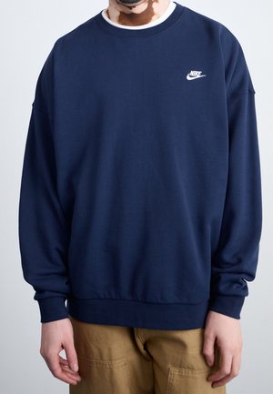 Mężczyzna w granatowej bluzie Nike i khaki spodniach, stojący na jednolitym jasnym tle.
