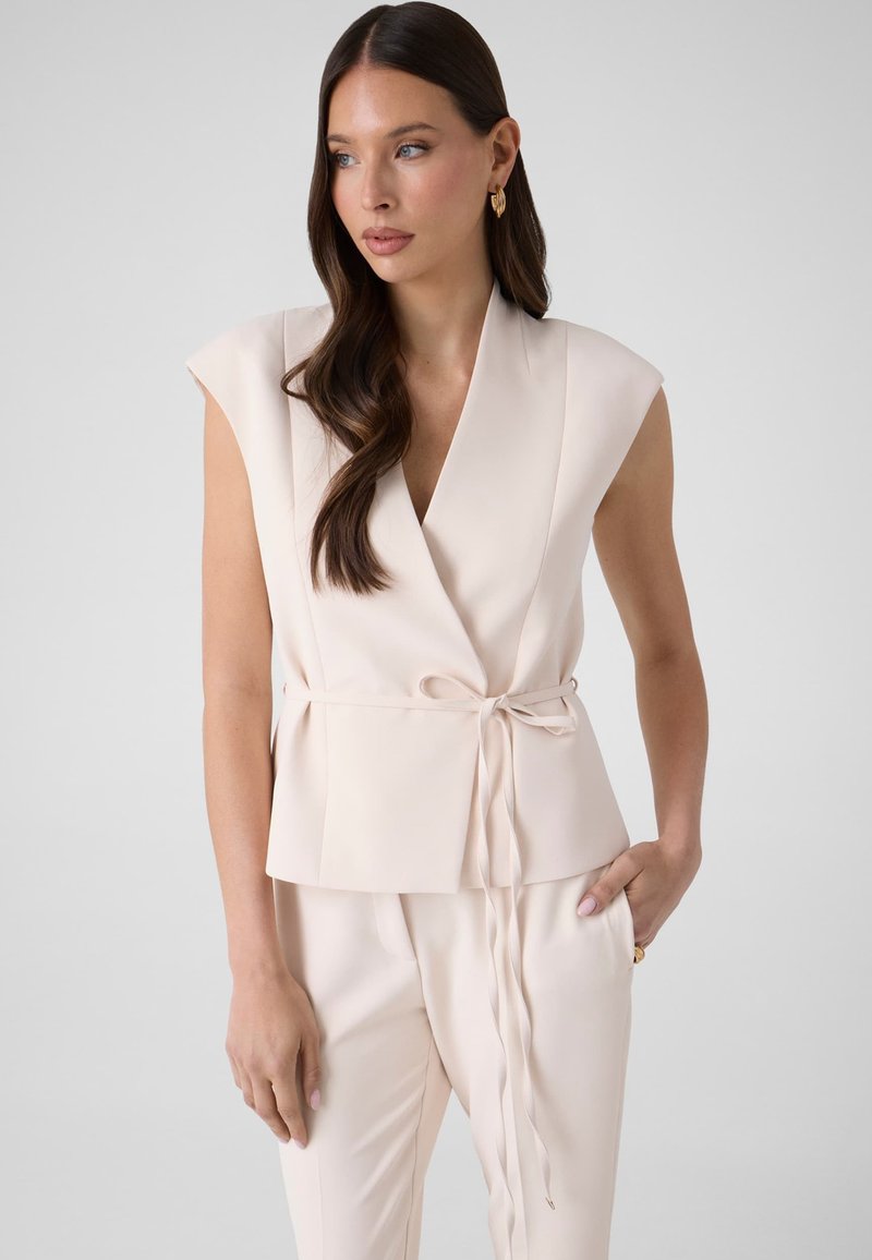 Femme aux longs cheveux bruns portant un chemisier beige sans manches avec ceinture nouée et pantalon assorti, regardant sur le côté devant un fond uni.