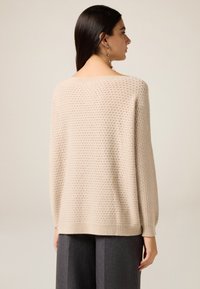 Beżowy, dziergany sweter o luźnym kroju, z teksturowanym wzorem i ściągaczami przy rękawach. Noszony z szarymi, wysokim stanem spodniami.