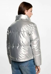 Srebrna puffer jakna z visokim ovratnikom, prešita zasnova in sijoč zaključek. Ima elastične manšete in rob, v kombinaciji z modrimi jeans hlačami.