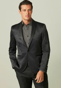Schwarze Satin-Blazer mit Reverskragen, einreihigem Knopfverschluss und zwei Fronttaschen, getragen über einem gemusterten schwarz-weißen Hemd.