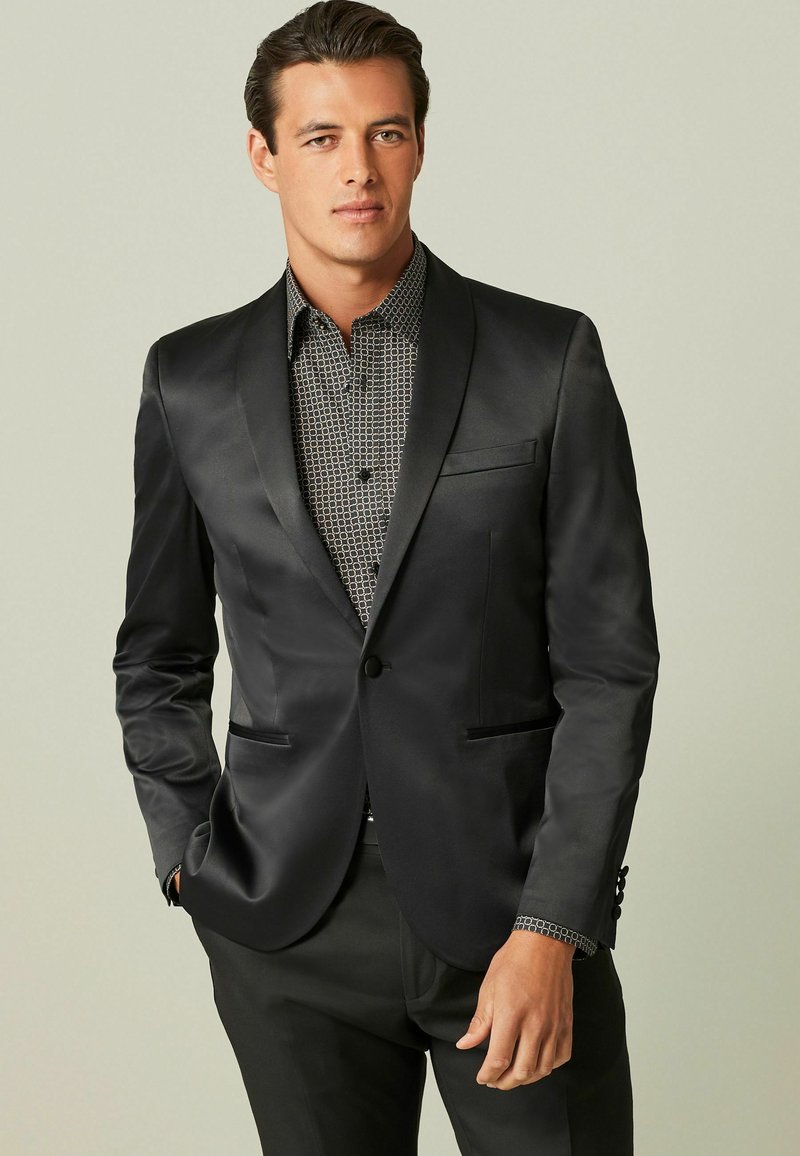 Schwarze Satin-Blazer mit Reverskragen, einreihigem Knopfverschluss und zwei Fronttaschen, getragen über einem gemusterten schwarz-weißen Hemd.