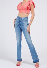 Jean denim bleu clair évasé avec ourlet effiloché, associé à un court haut rose avec manches volantées et détails en œillets. Sandales à talons roses.