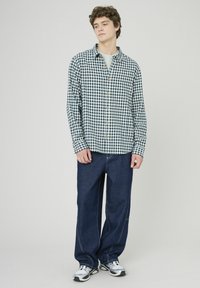 Chemise à carreaux verte et marine avec fermeture à boutons, associée à un jean en denim sombre ample. Le modèle se tient avec les mains dans les poches.