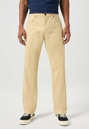 Pantaloni di cotone color crema con un design a gamba dritta, caratterizzati da tasche frontali, chiusura con bottone e una texture sottile. Abbinati a sneaker nere.
