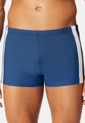 Männlicher Oberkörper trägt enganliegende blaue Badeshorts mit weißen vertikalen Seitenstreifen vor einem einfarbigen hellen Hintergrund.
