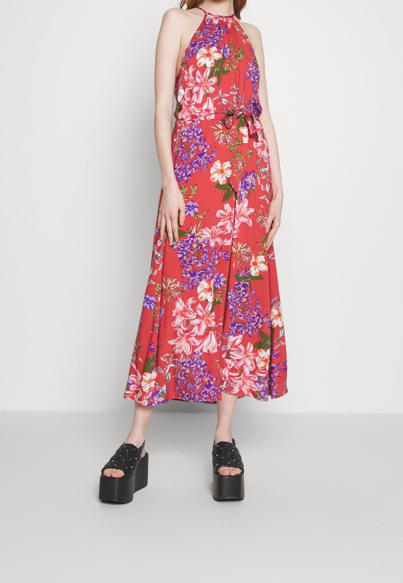 Femme portant une robe rouge à fleurs sans manches avec un col halter et des sandales compensées noires, debout devant un fond uni.