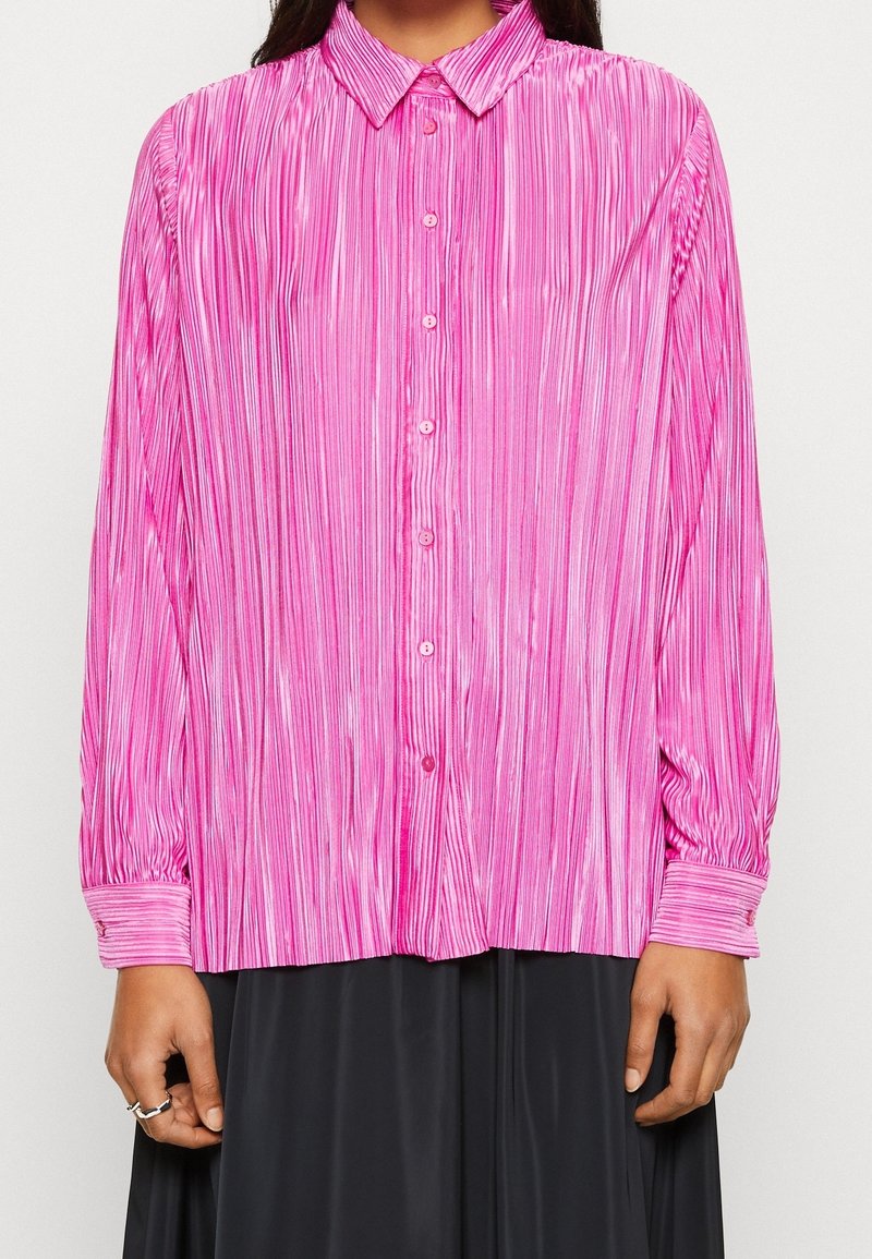 Camicia rosa a maniche lunghe con piastre verticali, abbottonatura frontale e colletto classico; presenta una texture liscia e lucida e maniche con polsini.