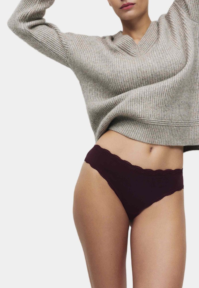 Tanga vínové barvy s krajkovým lemem a texturovaným designem, doplněná světle šedým pleteným svetrem. Zaměřte se na látku a střih.