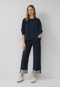 Donkerblauwe rits-sweater met een ronde hals, gecombineerd met brede donkere spijkerbroeken met opgerolde pijpen en witte sneakers.