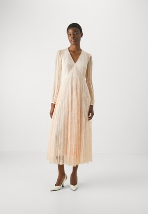 Long, blush-colored dress with a textured lace bodice, sheer long sleeves, and a flowing pleated skirt. Paired with white heeled shoes.