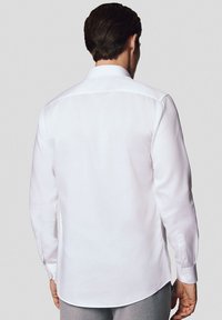 Biała koszula z długim rękawem, zapinana na guziki, z prostym kołnierzykiem, dopasowanym krojem i gładką fakturą; posiada guziki przy mankietach i zaokrąglony dół.