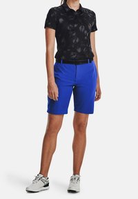 Vuxen klädd i svart mönstrad kortärmad polo, ljusblå knäkorta shorts med svart bälte och vita träningsskor, stående på vit bakgrund.