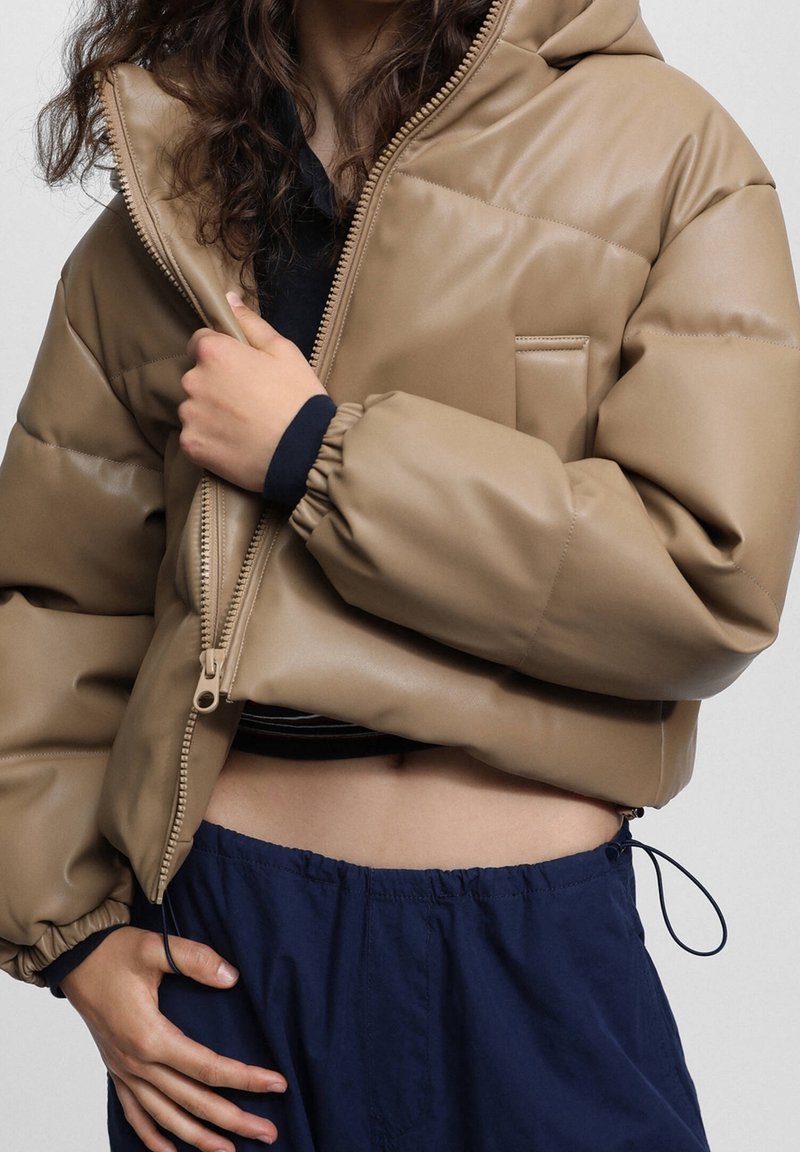Personne portant une doudoune courte beige avec des poignets élastiques, partiellement dézippée, dévoilant un haut noir et un pantalon bleu marine à cordon.