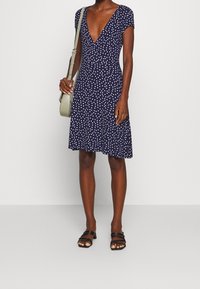 Kvinna i en knälång marinblå klänning med prickar, korta ärmar, svarta klacksandaler och en ljusgrön axelväska.