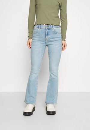 Personne portant un jean flare taille haute bleu clair, un haut côtelé manches longues vert olive et des bottines à plateforme blanches, debout devant un fond uni.