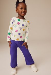 Ribbade långärmade topp med flerfärgade molnmönster; lila texturerade byxor med vågig fåll; vita sneakers fulländar outfiten.