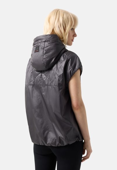 Lyshåret kvinde iført en ærmeløs mørkegrå hættetrøje, vist bagfra mod en lysegrå baggrund.