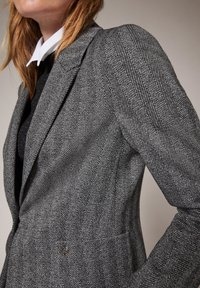 Blazer à motif pied-de-poule en gris et blanc, doté d'une épaule structurée, d'une coupe ajustée, et d'un petit bouton métallique.