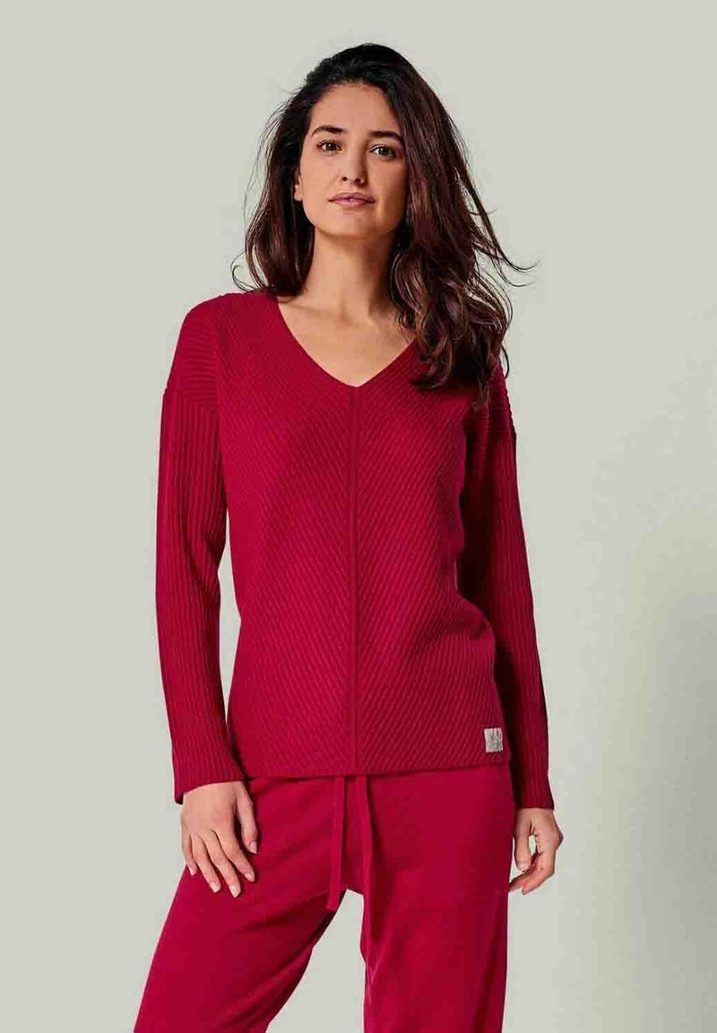 Roter gerippter Strickpullover mit langen Ärmeln und V-Ausschnitt, kombiniert mit passenden Hosen. Er zeichnet sich durch eine vertikale Nahtdetails und ein kleines Logotag am Saum aus.