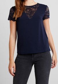 Navy blue top with short lace sleeves and a V-shaped lace inset. The fabric is smooth, with a relaxed fit and a rounded neckline.