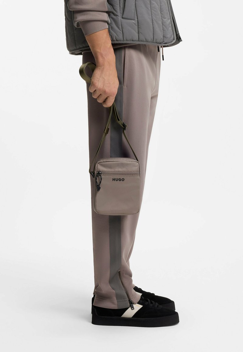 Personne portant un pantalon taupe avec des bandes latérales foncées et des baskets noires, tenant un sac bandoulière HUGO taupe par la sangle.