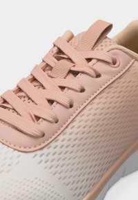 Κοντινή λήψη ενός ροζ και μπεζ αθλητικού παπουτσιού με υφή υφάσματος, επίπεδα κορδόνια και θηλιά έλξης στη γλώσσα.