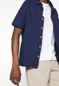 Kortärmad, marinblå knapp-up skjorta i lätt tyg. Har en krage och vita knappar, lager-på-lager över en vit t-shirt.