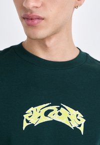 Green cotton t-shirt featuring a large yellow and white graphic print on the chest, round neckline, and short sleeves.