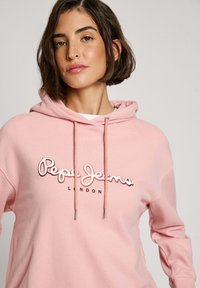 Felpa di cotone rosa con cappuccio regolabile, caratterizzata da un grande logo ricamato "Pepe Jeans London" in bianco e marrone scuro. Texture liscia.