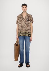 Camisa de manga corta con estampado de leopardo y logo negro, combinada con jeans azules y sandalias negras. Sosteniendo una bolsa de papel marrón.