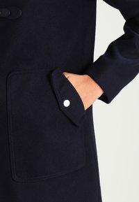 Mörkblå kappa i mjukt tyg med en stor ficka med tryckknapp. Ärmarna är skräddarsydda och åtsittande.