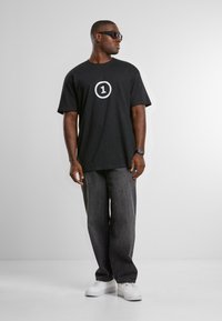 Svart bomull T-skjorte med en hvit sirkel og "1" grafikk, kombinert med grå denimbukser og hvite sneakers. Personen har på seg solbriller.