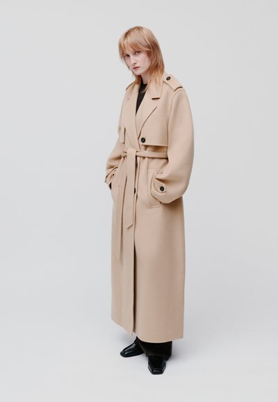 Cappotto lungo in misto lana beige con vita cintata. Presenta ampi rever, bottoni doppi e maniche oversized. Le scarpe nere completano l'outfit.