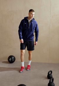 Marine blauw zip-up hoodie, zwarte shorts, witte sokken en rode sportschoenen. Twee kettlebells en een medicijnbal op een grijze vloer.