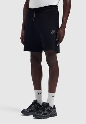 Man met zwarte sportkorte broek met trekkoord, witte Nike sportsokken en zwarte hardloopschoenen, staande tegen een effen achtergrond.