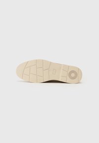 Suela de zapatilla beige con una superficie de goma texturizada. Presenta un patrón circular y una sección rectangular cerca del talón para mayor agarre.