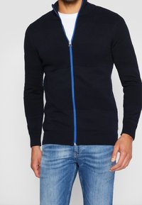 Marinblå stickad zip-up-tröja med texturerade horisontella ränder, kontrasterande blå dragkedja, hög krage samt ribbade muddar och fåll.