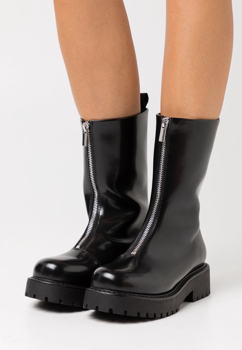 Bottes noires brillantes mi-mollet avec semelles épaisses et fermetures éclair argentées à l'avant portées sur des jambes nues sur un fond uni.